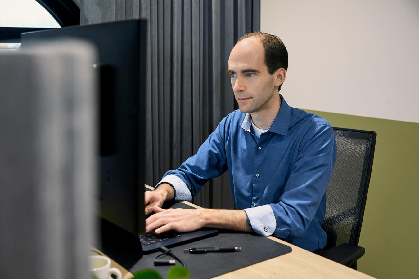 Werknemer in blauwe blouse achter computer in het kantoor van NextUp. Hij kijkt serieus naar zijn laptop.